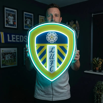 A man holding a large shield-shaped Leeds United FC football club crest LED neon sign featuring the Yorkshire White Rose and LUFC script glowing in vibrant yellow and cyan blue light