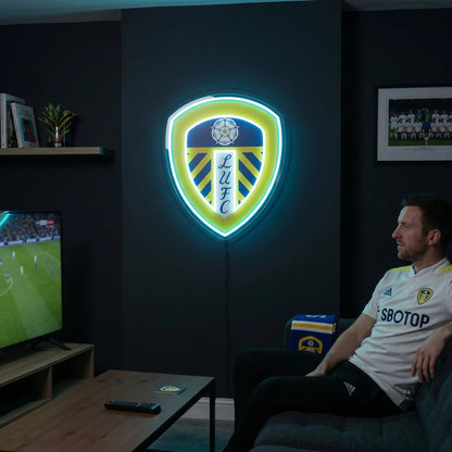 A man holding a large shield-shaped Leeds United FC football club crest LED neon sign featuring the Yorkshire White Rose and LUFC script glowing in vibrant yellow and cyan blue light