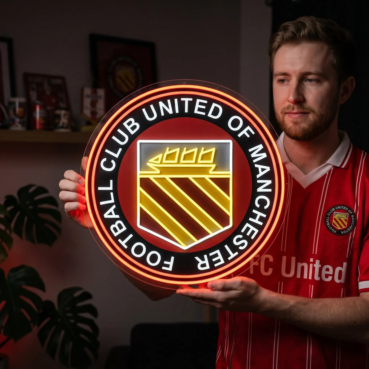 A man holding a large circular FC United of Manchester football club crest LED neon sign featuring the traditional ship and three diagonal stripes shield glowing in warm yellow, white, and red light