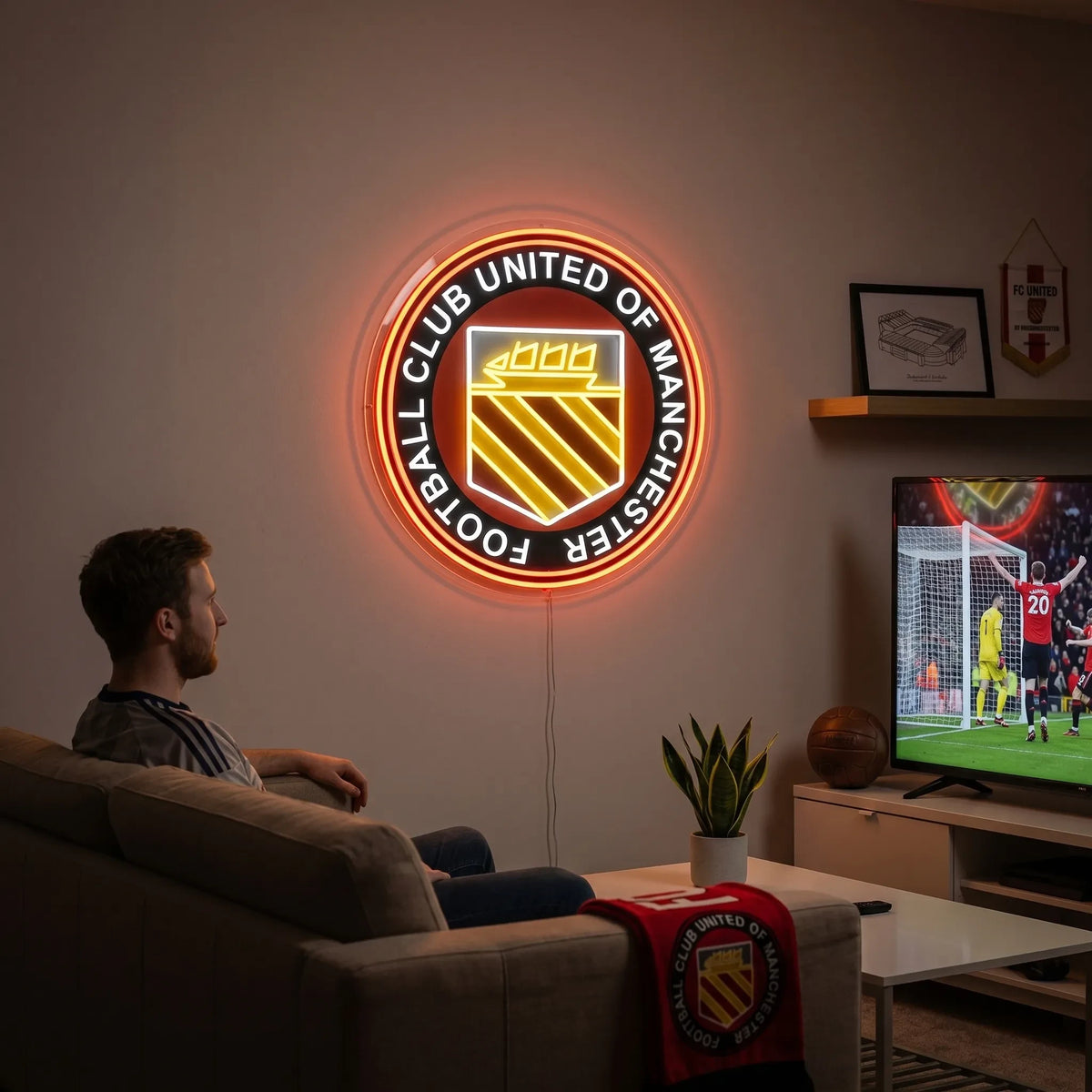 A man holding a large circular FC United of Manchester football club crest LED neon sign featuring the traditional ship and three diagonal stripes shield glowing in warm yellow, white, and red light