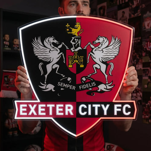 A man holding a shield-shaped Exeter City FC football club crest LED neon sign with a red and black split background, featuring the two winged horses, a castle emblem, and the Semper Fidelis motto glowing in bright white and red light