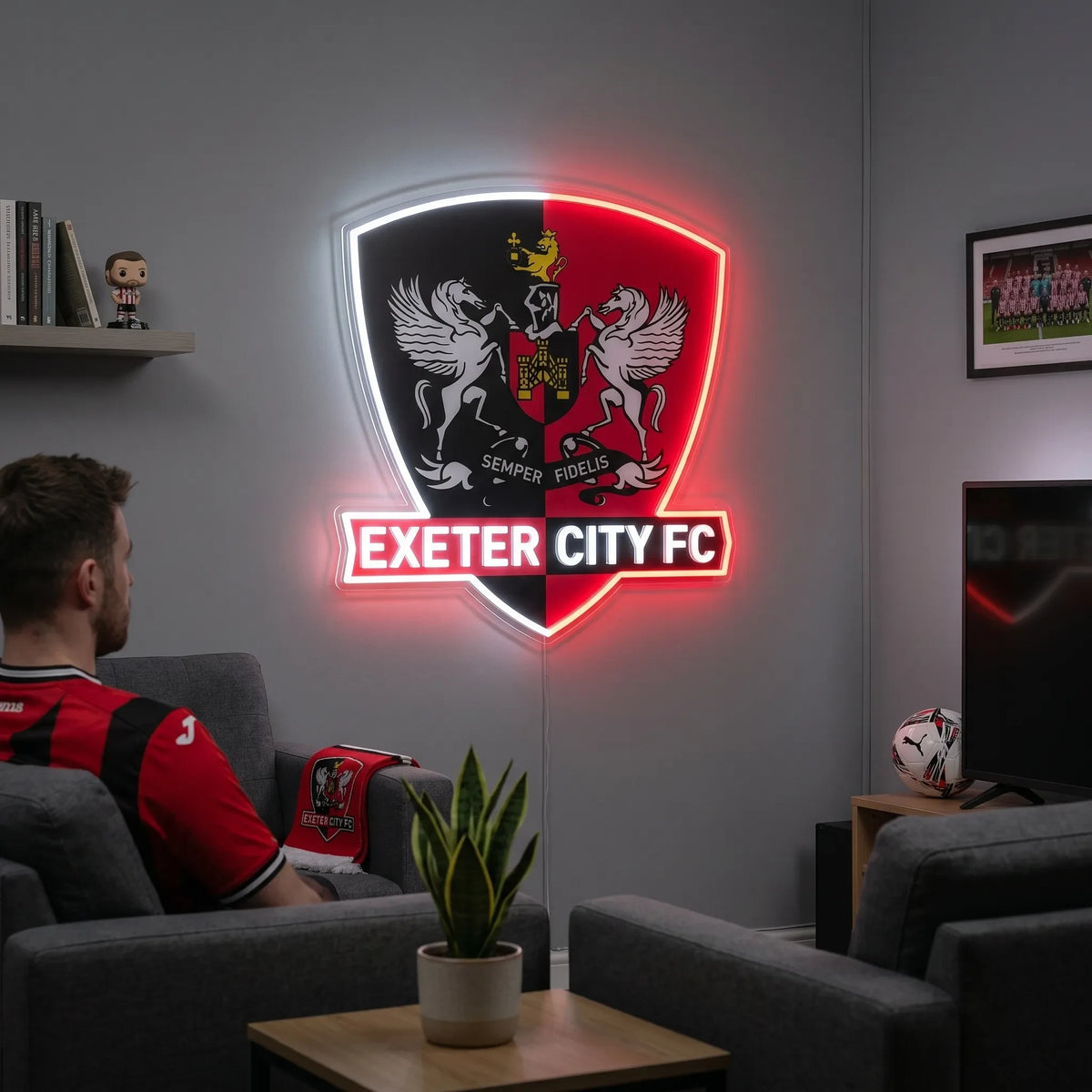 A man holding a shield-shaped Exeter City FC football club crest LED neon sign with a red and black split background, featuring the two winged horses, a castle emblem, and the Semper Fidelis motto glowing in bright white and red light