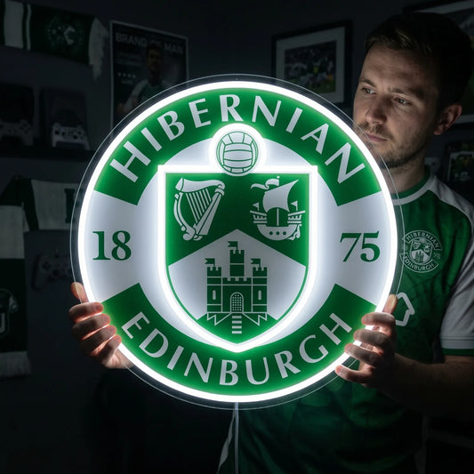 A man holding a large circular Hibernian FC Edinburgh football club crest LED neon sign featuring the harp, ship, and Edinburgh Castle shield glowing in vibrant green and white light