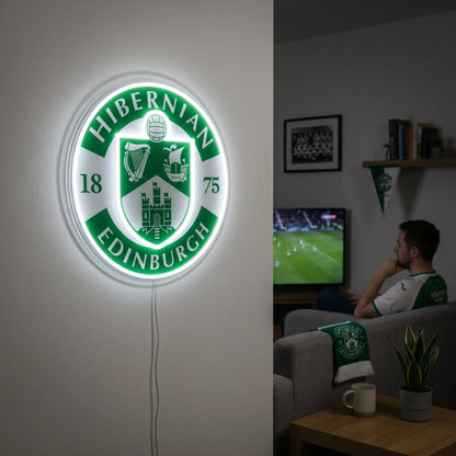 A man holding a large circular Hibernian FC Edinburgh football club crest LED neon sign featuring the harp, ship, and Edinburgh Castle shield glowing in vibrant green and white light