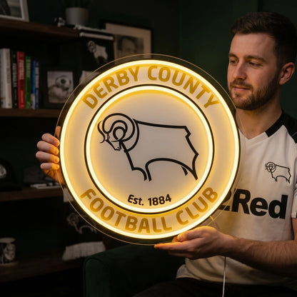 A man holding a large circular Derby County Football Club crest LED neon sign featuring the iconic Ram logo glowing in warm yellow light