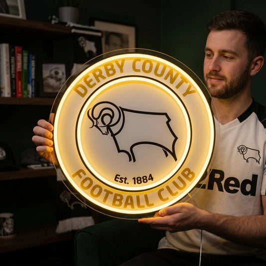 A man holding a large circular Derby County Football Club crest LED neon sign featuring the iconic Ram logo glowing in warm yellow light