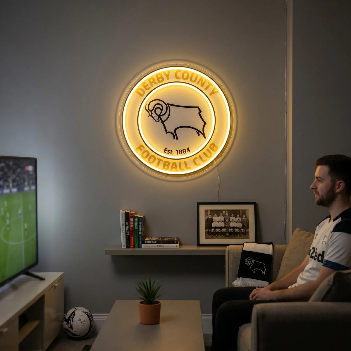 A man holding a large circular Derby County Football Club crest LED neon sign featuring the iconic Ram logo glowing in warm yellow light