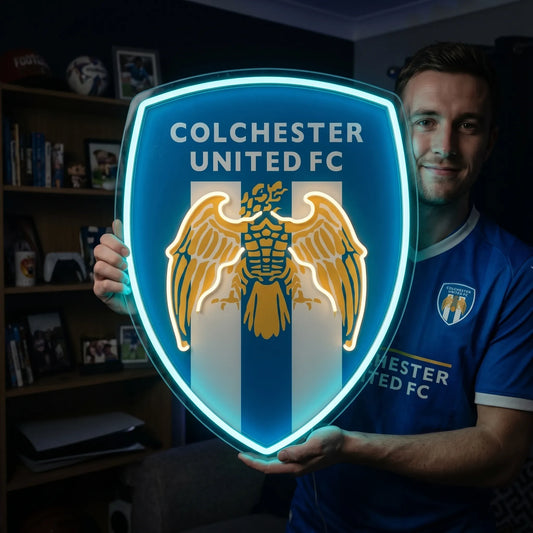 A man holding a shield-shaped Colchester United FC football club crest LED neon sign featuring the Roman eagle logo glowing in blue and warm yellow light