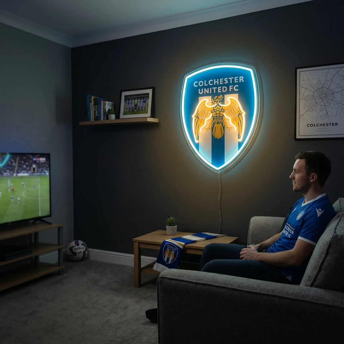 A man holding a shield-shaped Colchester United FC football club crest LED neon sign featuring the Roman eagle logo glowing in blue and warm yellow light