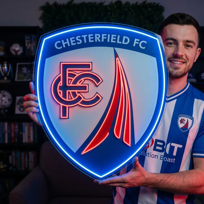 A man smiling and holding a large shield-shaped Chesterfield FC football club crest LED neon sign featuring the CFC monogram and the famous crooked spire design glowing in blue and red light