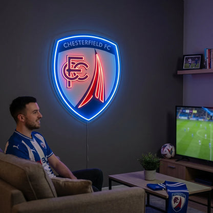 A man smiling and holding a large shield-shaped Chesterfield FC football club crest LED neon sign featuring the CFC monogram and the famous crooked spire design glowing in blue and red light