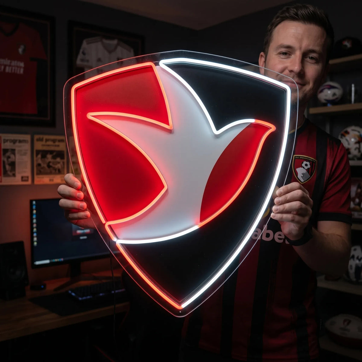 A man holding a shield-shaped AFC Bournemouth football club crest LED neon sign featuring the iconic stylized profile of Dickie Dowsett heading a football glowing in red and white light