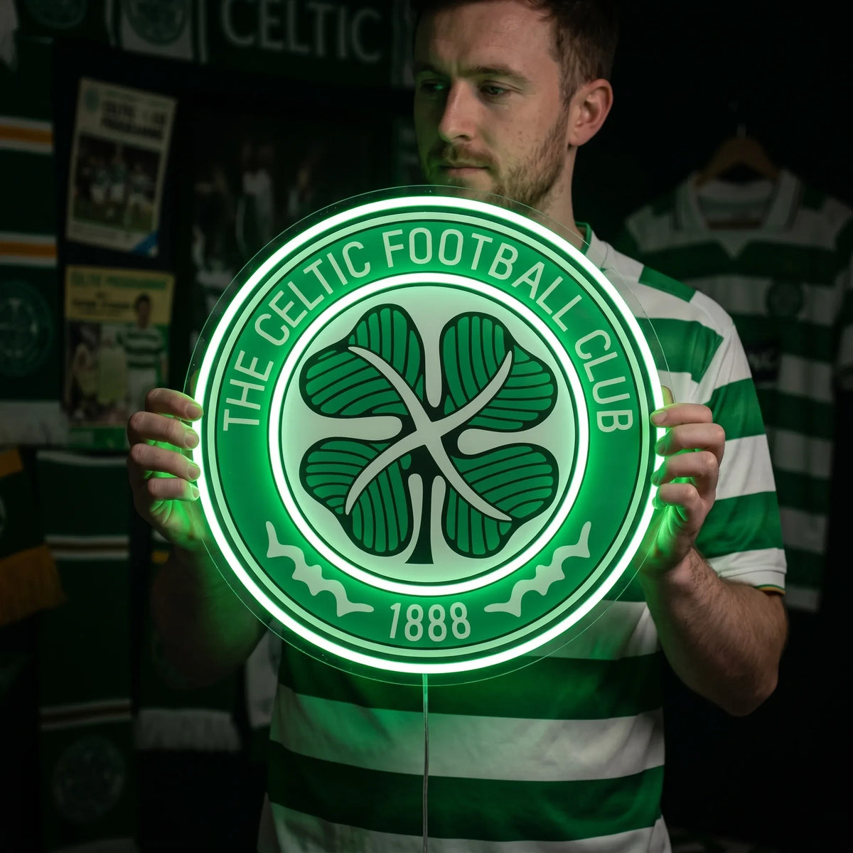 A man holding a circular The Celtic Football Club crest LED neon sign featuring the traditional four-leaf clover logo glowing in a vibrant green light