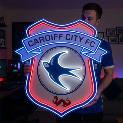 A man holding a shield-shaped Cardiff City FC football club crest LED neon sign featuring the iconic bluebird and red dragon glowing in blue, white, and red light