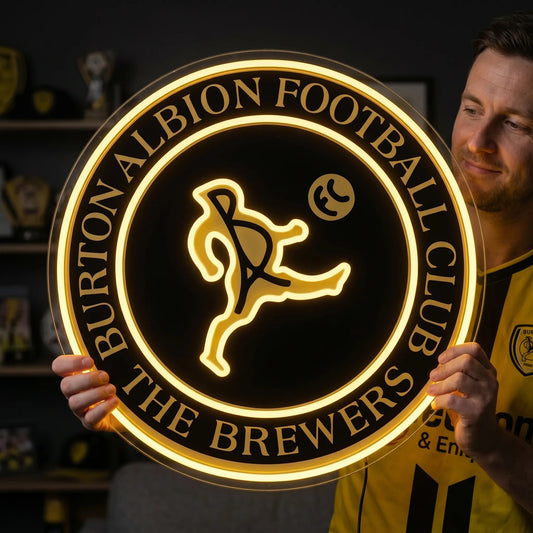 A man holding a large circular Burton Albion Football Club crest LED neon sign featuring the stylized kicker logo and The Brewers nickname glowing in warm yellow light