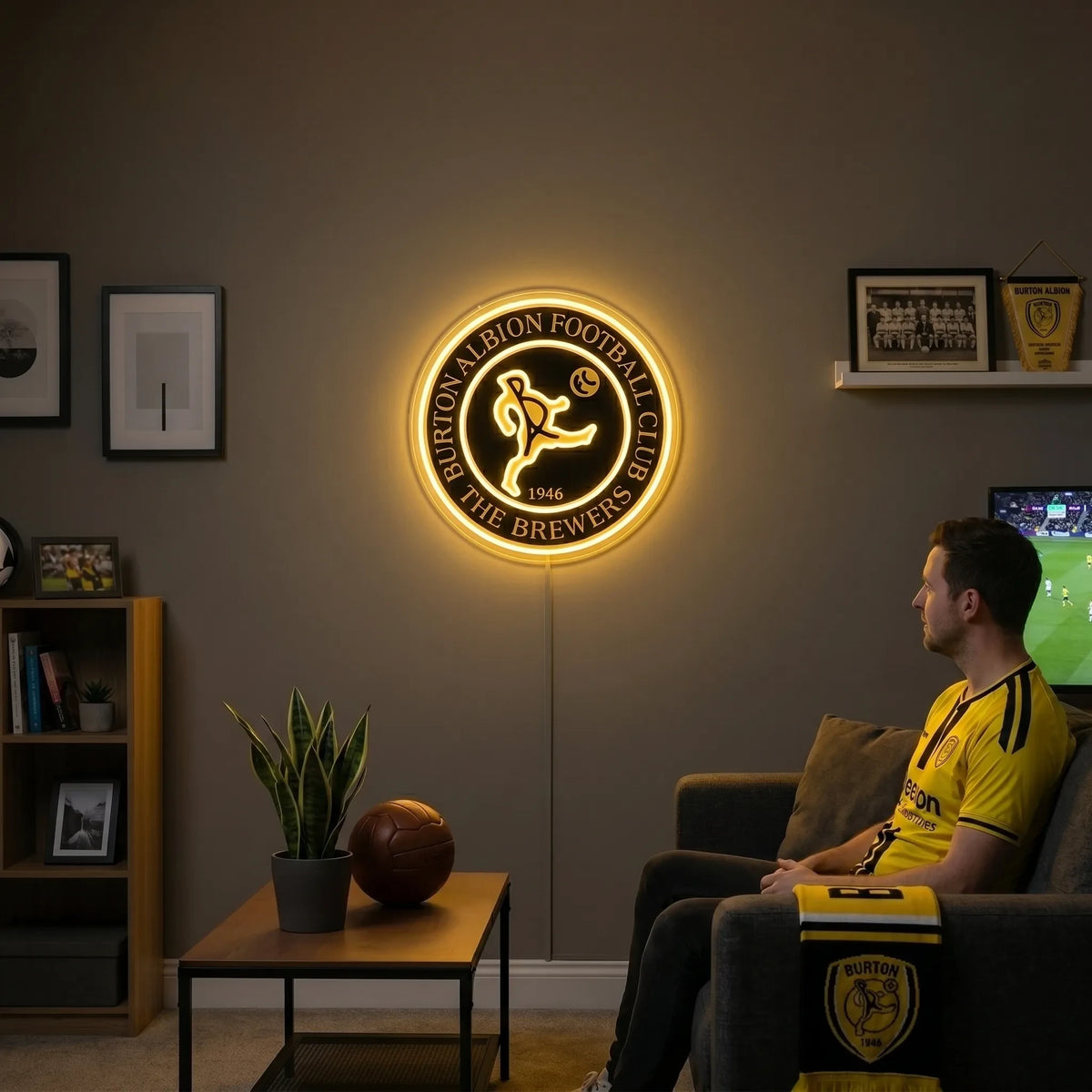 A man holding a large circular Burton Albion Football Club crest LED neon sign featuring the stylized kicker logo and The Brewers nickname glowing in warm yellow light