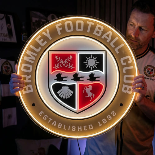 A man holding a large circular Bromley Football Club crest LED neon sign featuring the quad-divided shield with a sun, horse, and ravens glowing in warm yellow, white, and red light