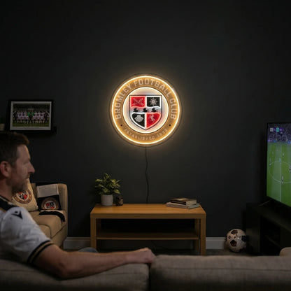 A man holding a large circular Bromley Football Club crest LED neon sign featuring the quad-divided shield with a sun, horse, and ravens glowing in warm yellow, white, and red light