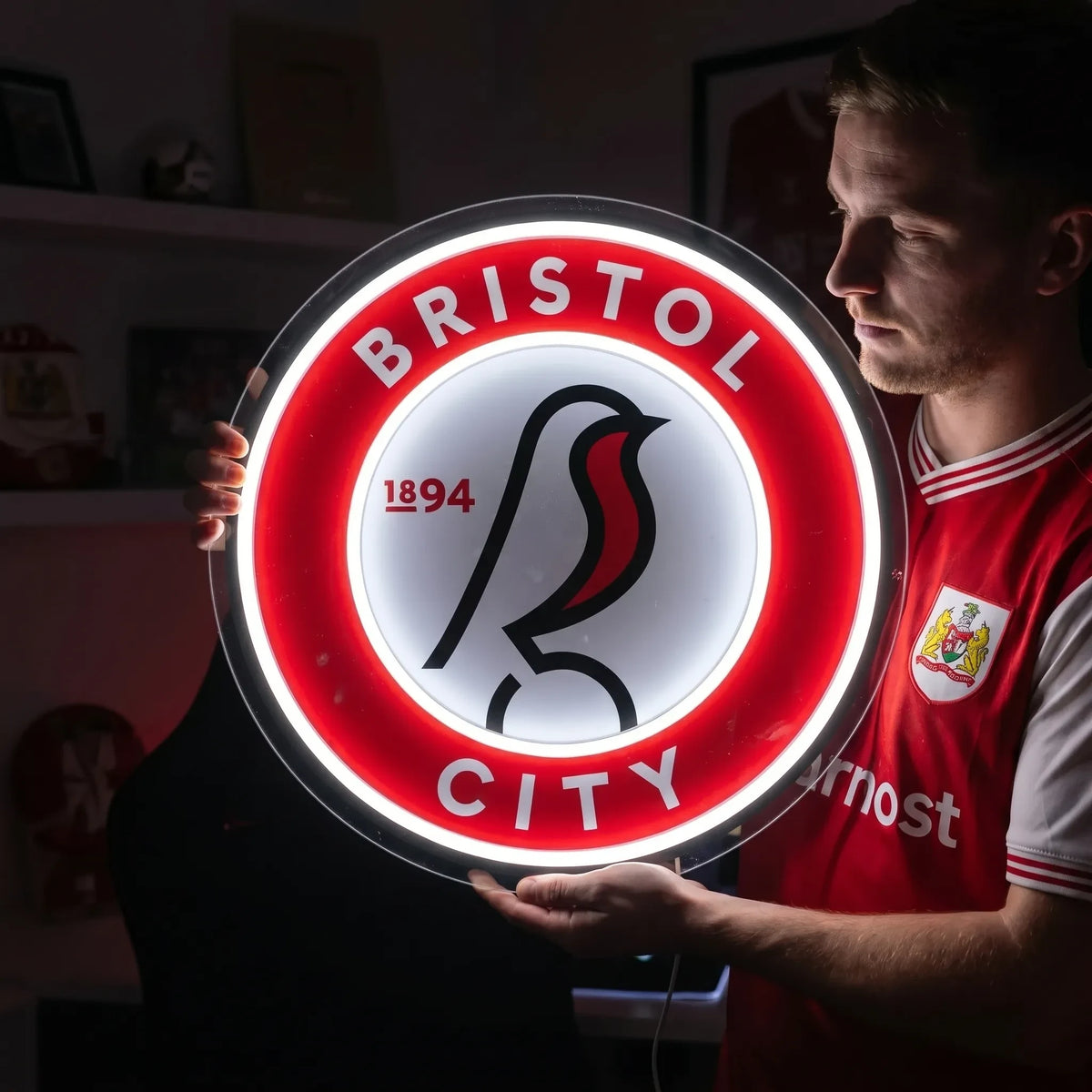 A man holding a circular Bristol City FC football club crest LED neon sign featuring the robin bird logo glowing in bright white and red light