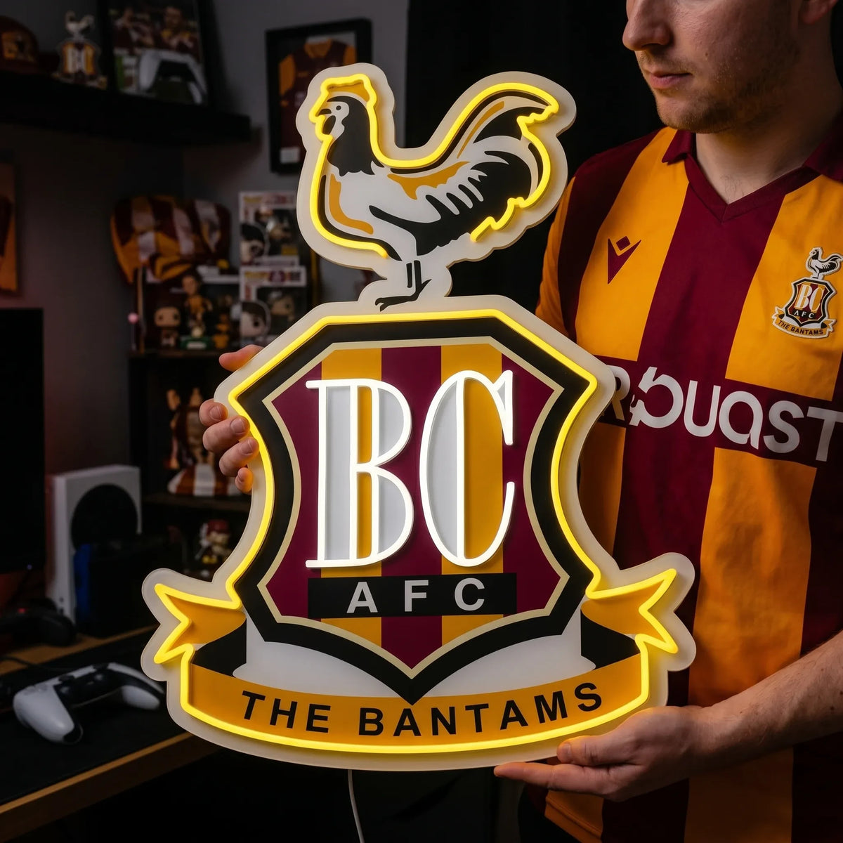 A man holding a custom Bradford City AFC football club crest LED neon sign featuring the BC initials, a bantam rooster on top, and The Bantams ribbon glowing in warm yellow and white light