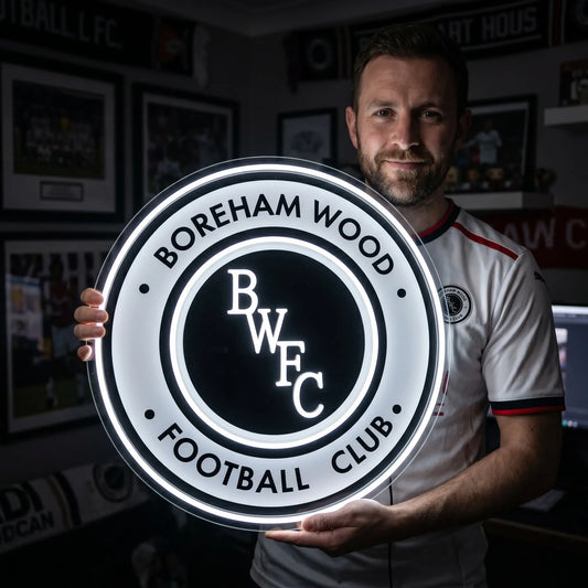 A man holding a large circular Boreham Wood FC football club crest LED neon sign with BWFC initials glowing in bright white light
