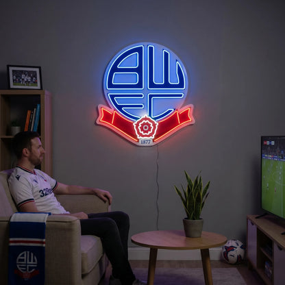 A man holding a circular Bolton Wanderers FC football club crest LED neon sign featuring the BWFC initials and the Red Rose of Lancashire ribbon glowing in blue and red light