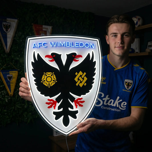 A man holding a shield-shaped AFC Wimbledon football club crest LED neon sign featuring the double-headed eagle logo glowing in a bright white and blue light