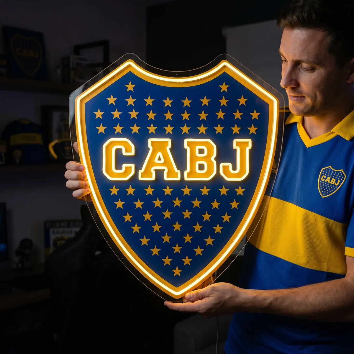 A shield-shaped Boca Juniors CABJ football club crest LED neon sign glowing in bright yellow light mounted on a living room wall above a TV setup with a fan watching from a sofa