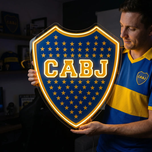 A shield-shaped Boca Juniors CABJ football club crest LED neon sign glowing in bright yellow light mounted on a living room wall above a TV setup with a fan watching from a sofa