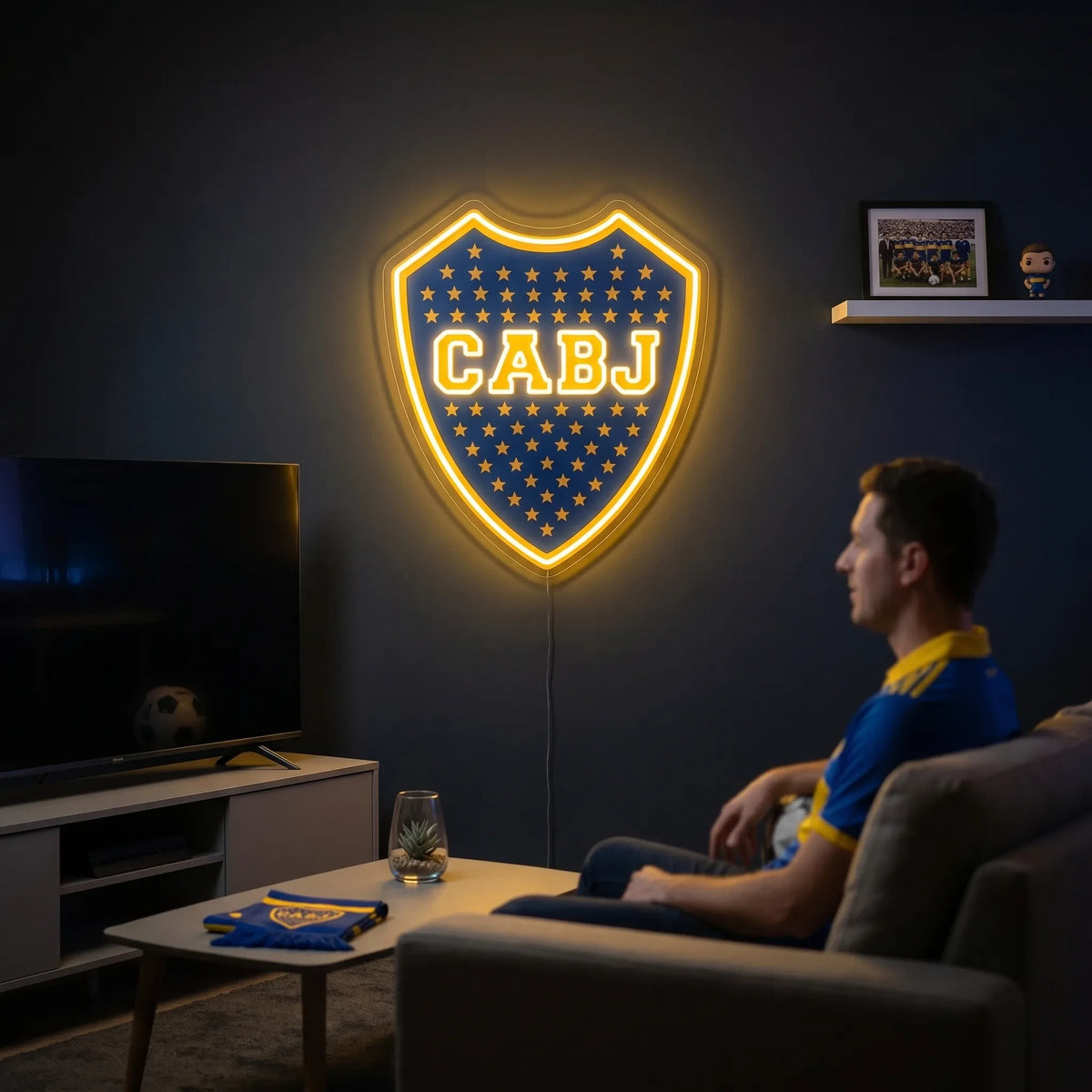 A shield-shaped Boca Juniors CABJ football club crest LED neon sign glowing in bright yellow light mounted on a living room wall above a TV setup with a fan watching from a sofa
