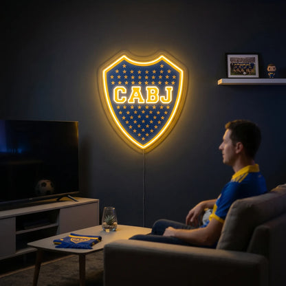 A shield-shaped Boca Juniors CABJ football club crest LED neon sign glowing in bright yellow light mounted on a living room wall above a TV setup with a fan watching from a sofa