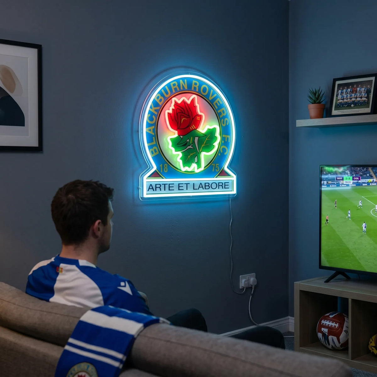 A man holding a Blackburn Rovers FC football club crest LED neon sign featuring the Red Rose of Lancaster and Latin motto Arte et Labore glowing in red, green, and blue light