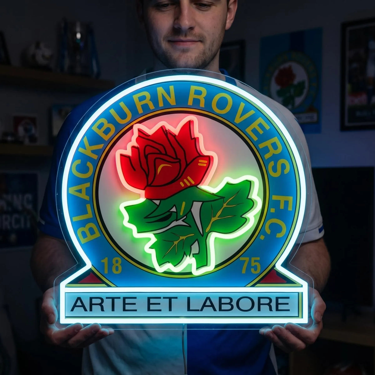 A man holding a Blackburn Rovers FC football club crest LED neon sign featuring the Red Rose of Lancaster and Latin motto Arte et Labore glowing in red, green, and blue light