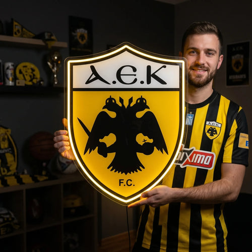 A smiling man holding a large shield-shaped AEK Athens FC football club crest LED neon sign featuring the black double-headed eagle and A.E.K. F.C. lettering glowing in a vibrant yellow light