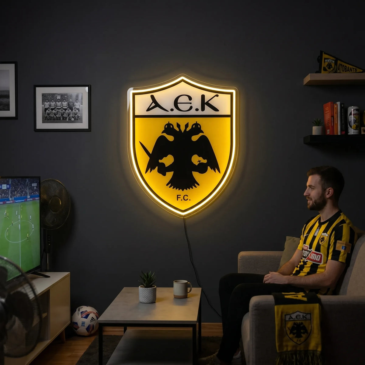 A smiling man holding a large shield-shaped AEK Athens FC football club crest LED neon sign featuring the black double-headed eagle and A.E.K. F.C. lettering glowing in a vibrant yellow light