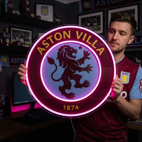 A man holding a circular Aston Villa FC football club crest LED neon sign featuring the lion logo glowing in a vibrant pinkish purple light