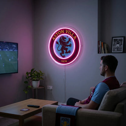 A man holding a circular Aston Villa FC football club crest LED neon sign featuring the lion logo glowing in a vibrant pinkish purple light