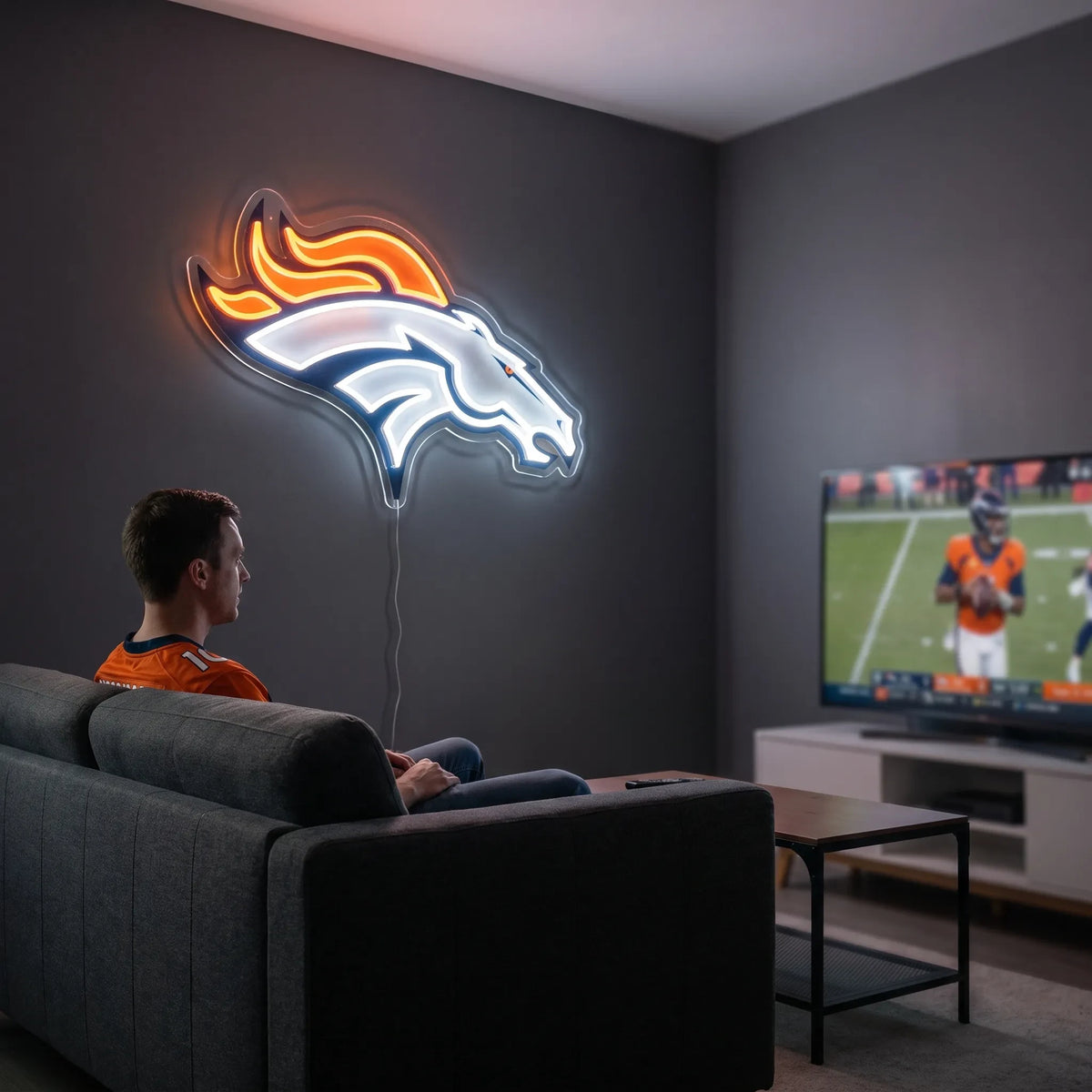 A man holding a large custom-shaped Denver Broncos NFL football team logo LED neon sign featuring the white bronco horse head profile with a bright orange flaming mane glowing in vibrant orange and crisp white light