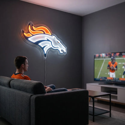 A man holding a large custom-shaped Denver Broncos NFL football team logo LED neon sign featuring the white bronco horse head profile with a bright orange flaming mane glowing in vibrant orange and crisp white light