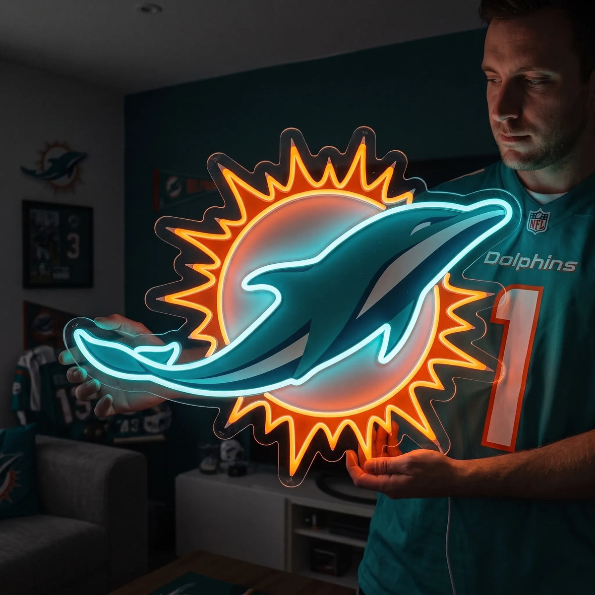 A man holding a large custom-shaped Miami Dolphins NFL football team logo LED neon sign featuring the leaping teal dolphin silhouette in front of an orange sunburst design glowing in vibrant aqua blue and bright orange light