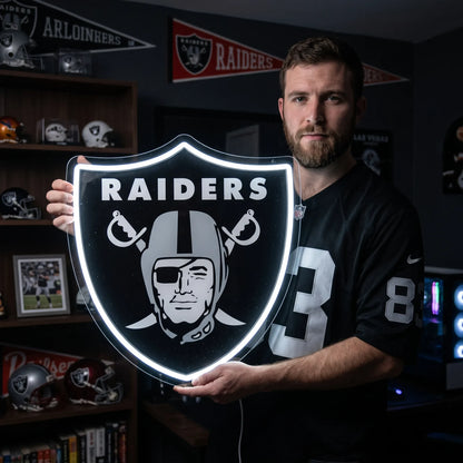 A man holding a large shield-shaped Las Vegas Raiders NFL football team logo LED neon sign featuring the iconic pirate head with an eyepatch wearing a football helmet over crossed cutlasses and the RAIDERS wordmark glowing in a bright white light