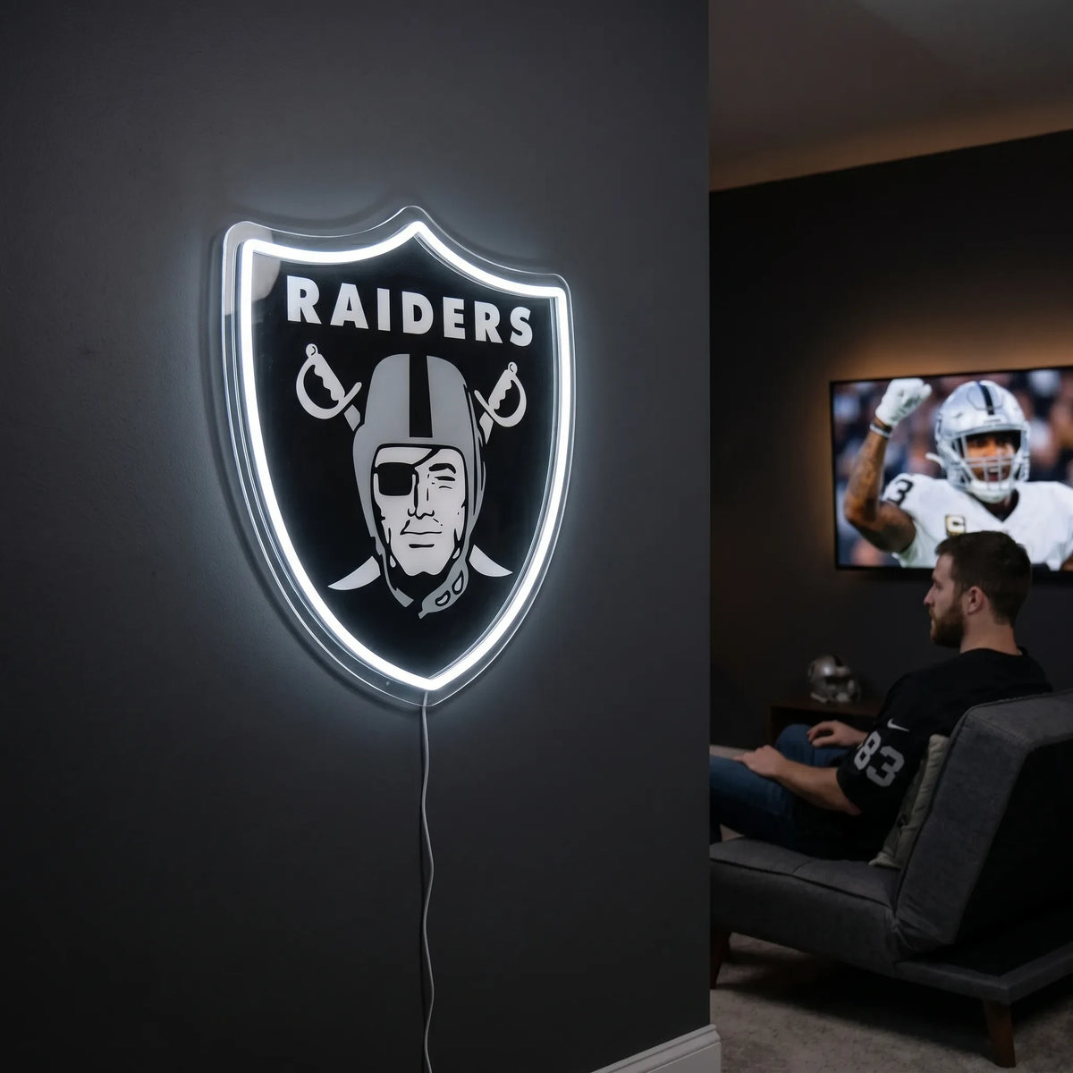 A man holding a large shield-shaped Las Vegas Raiders NFL football team logo LED neon sign featuring the iconic pirate head with an eyepatch wearing a football helmet over crossed cutlasses and the RAIDERS wordmark glowing in a bright white light