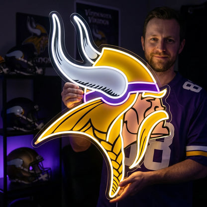 A smiling man holding a large custom-shaped Minnesota Vikings NFL football team logo LED neon sign featuring the Norseman head profile with a purple headband, a braided blonde mustache and hair, and white horns glowing in vibrant purple, gold yellow, and bright white light