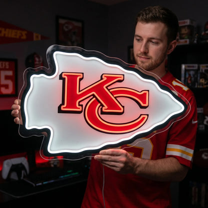 A man holding a large arrowhead-shaped Kansas City Chiefs NFL football team logo LED neon sign featuring the interlocking KC monogram in red with a bold black outline glowing in vibrant red and bright white light