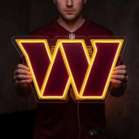 A man holding a large custom-shaped Washington Commanders NFL football team logo LED neon sign featuring the bold stylized letter W in burgundy outlined by a glowing vibrant yellow gold light