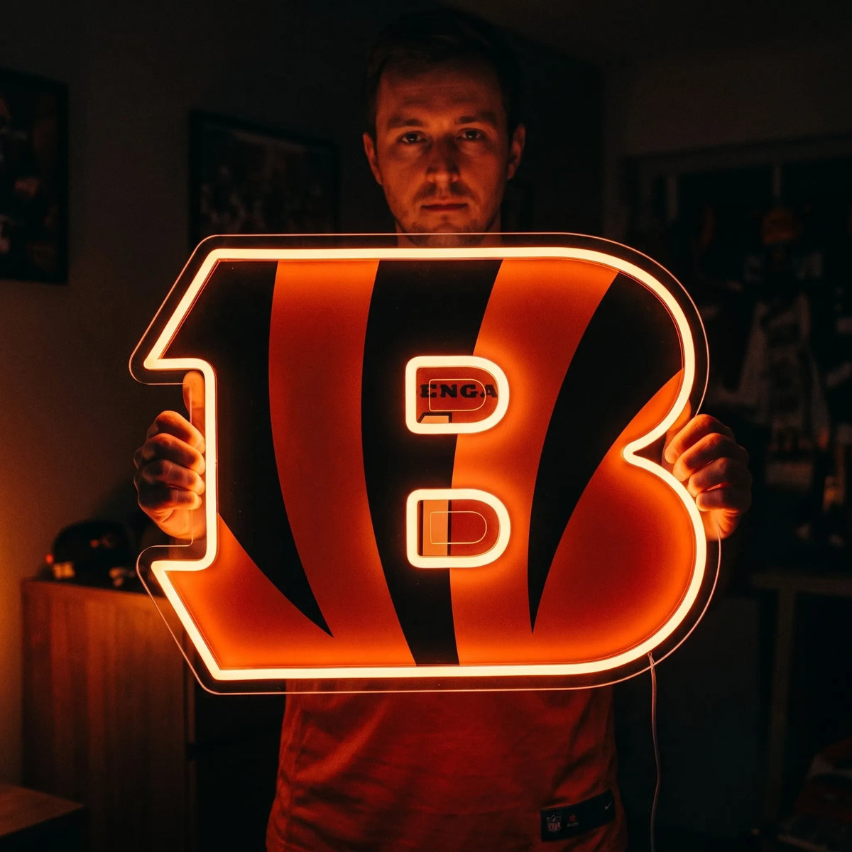 A man holding a large custom-shaped Cincinnati Bengals NFL football team logo LED neon sign featuring the bold letter B filled with black tiger stripes glowing in a vibrant bright orange light