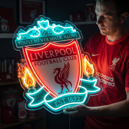 A person holding a glowing red and teal Liverpool FC LED neon sign featuring the Liver bird and "You'll Never Walk Alone" motto.