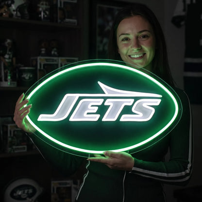 A man holding a large oval-shaped New York Jets NFL football team logo LED neon sign featuring the bold white JETS wordmark and airplane silhouette shape surrounded by a glowing vibrant green light border