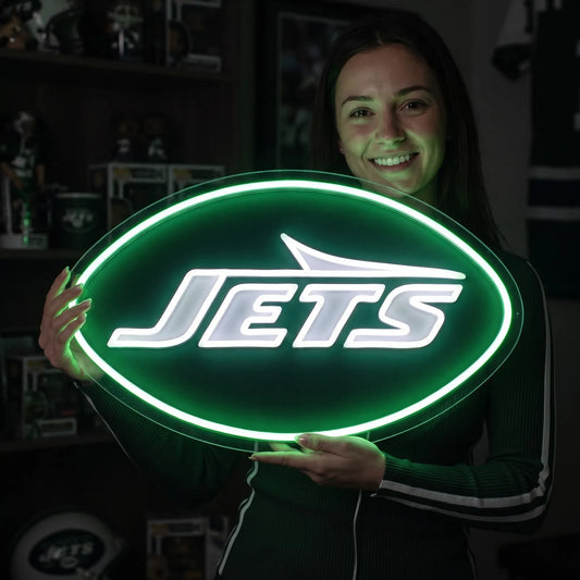 A man holding a large oval-shaped New York Jets NFL football team logo LED neon sign featuring the bold white JETS wordmark and airplane silhouette shape surrounded by a glowing vibrant green light border