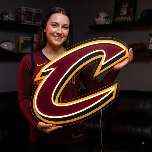 A man sitting at a gaming setup under a large glowing red and gold Cleveland Cavaliers NBA basketball team C logo LED neon sign mounted on a brick wall